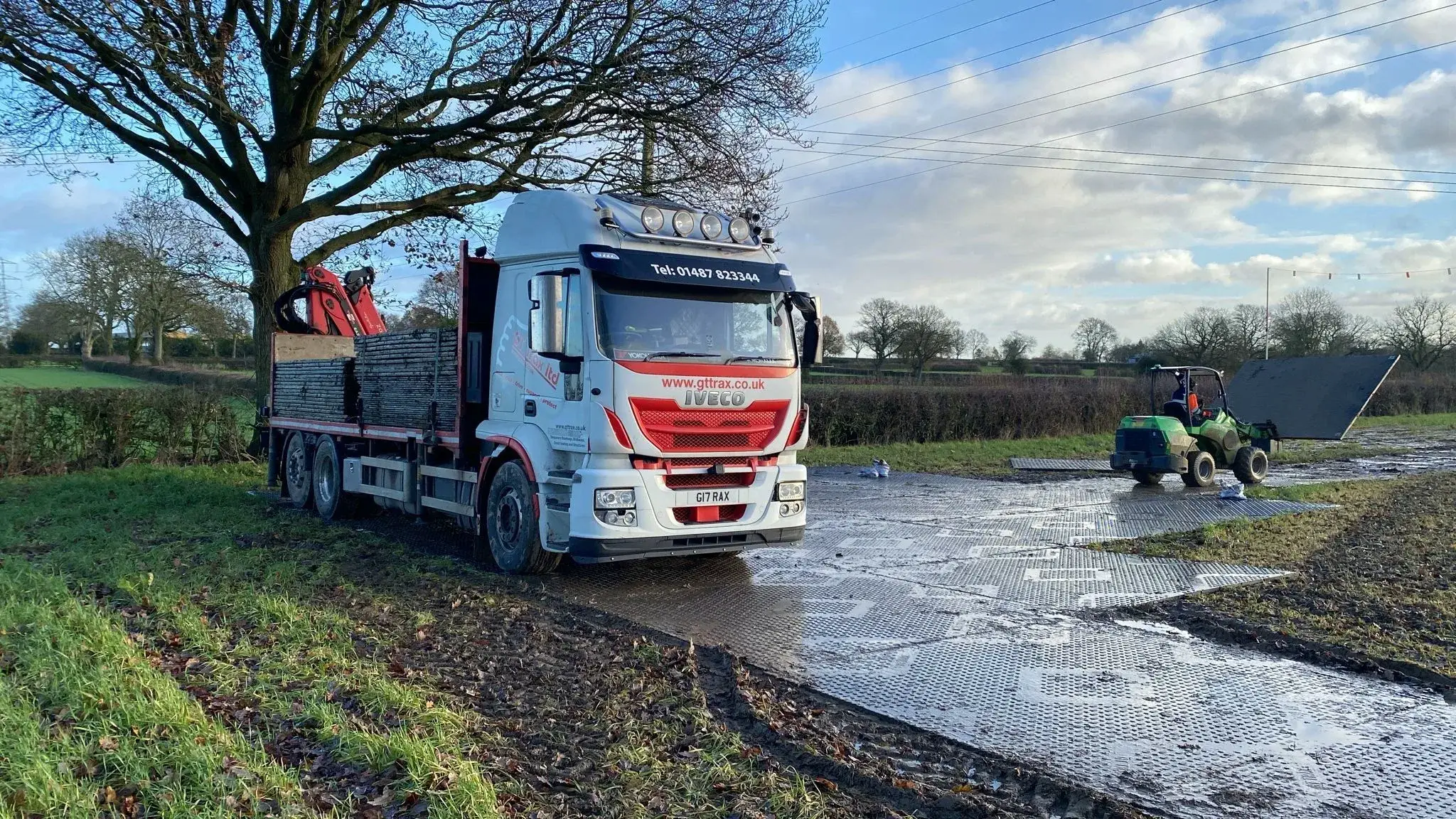 White truck with crane and green utility vehicle laying temporary PowerTrax 60 mats on a muddy field path.