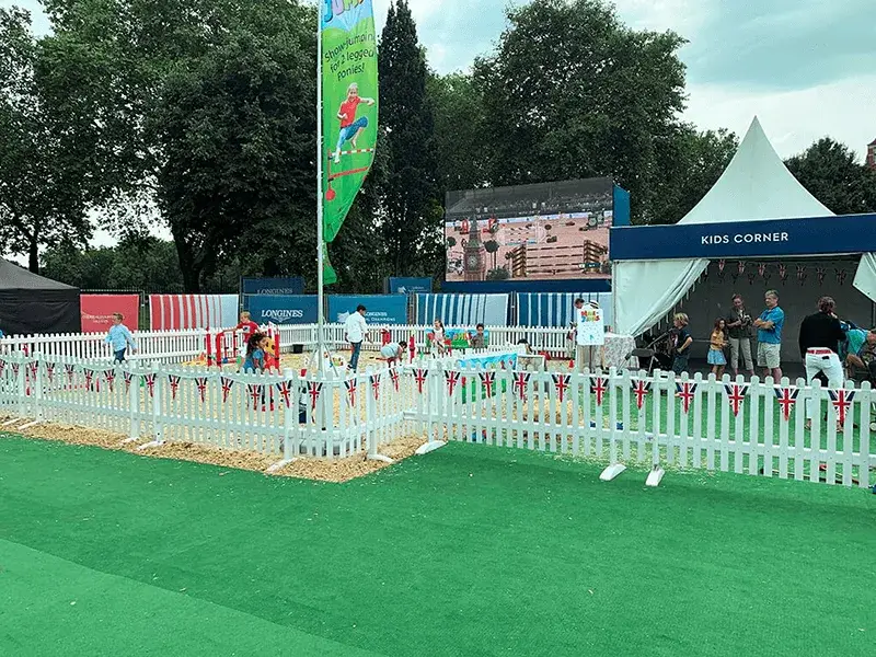Kids play in a picket fenced "Kids Corner" area with Union Jack bunting at an outdoor event, adults stand by. Large screen shows a competition behind the picket fencing.