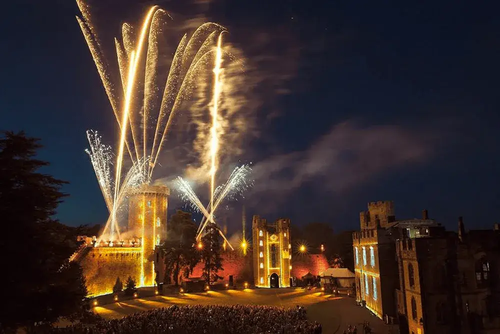 Golden fireworks streak above a brightly lit castle at night, with a large crowd gathered in the courtyard.