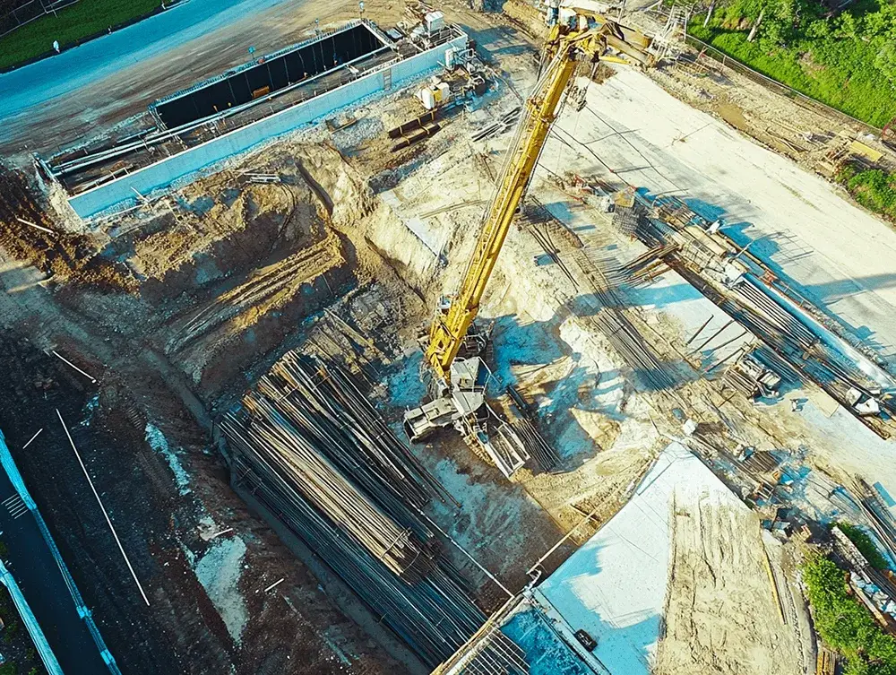 Aerial view of a large construction site with a yellow crane, excavated earth, stacks of pipes, and concrete structures.