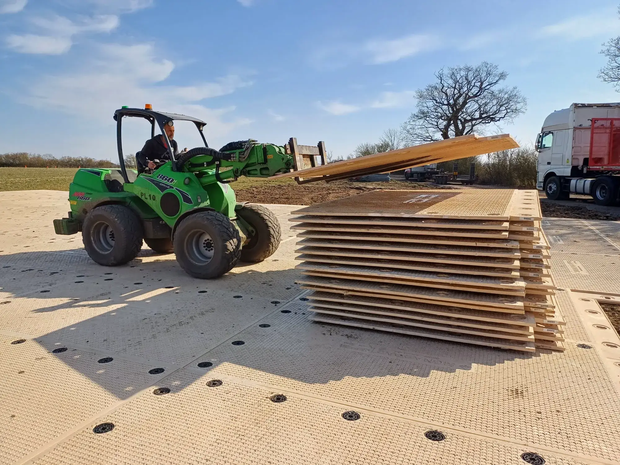 Green Avant loader lifts a panel, adding it to a stack of panels on temporary beige PowerTrax 60 ground mats under a clear sky.