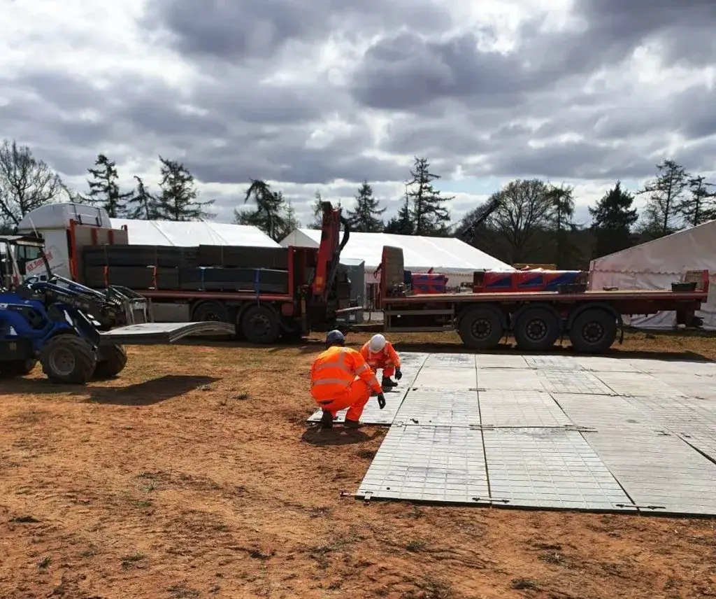 Two workers in high-vis orange lay gray PowerTrax 20 ground mats on dirt. A forklift and trucks with cargo are behind them, with white tents.