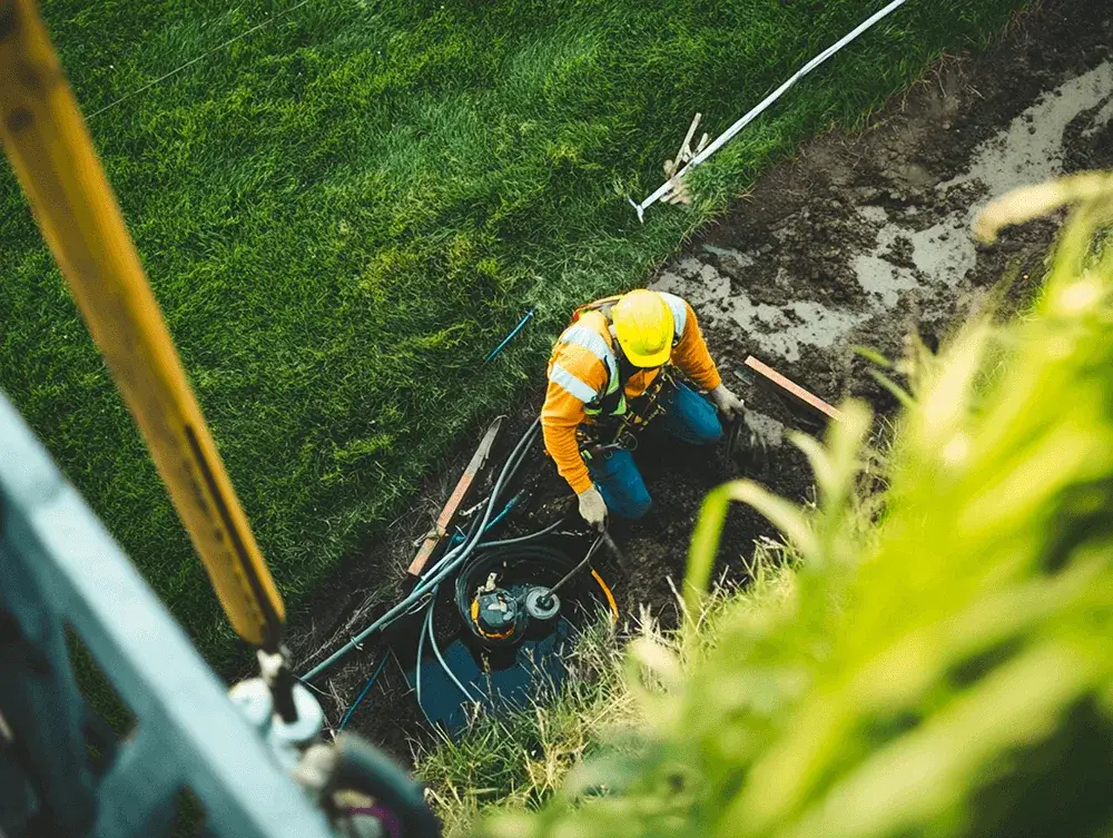 Overhead view of a worker in a hard hat and safety vest kneeling in a muddy trench, working with cables.