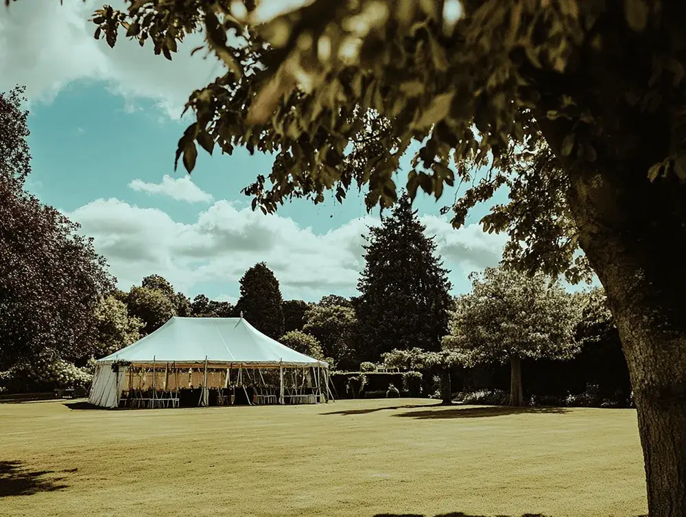 A large white event tent stands on a green lawn, surrounded by trees, under a partly cloudy blue sky. Leaves frame the top.
