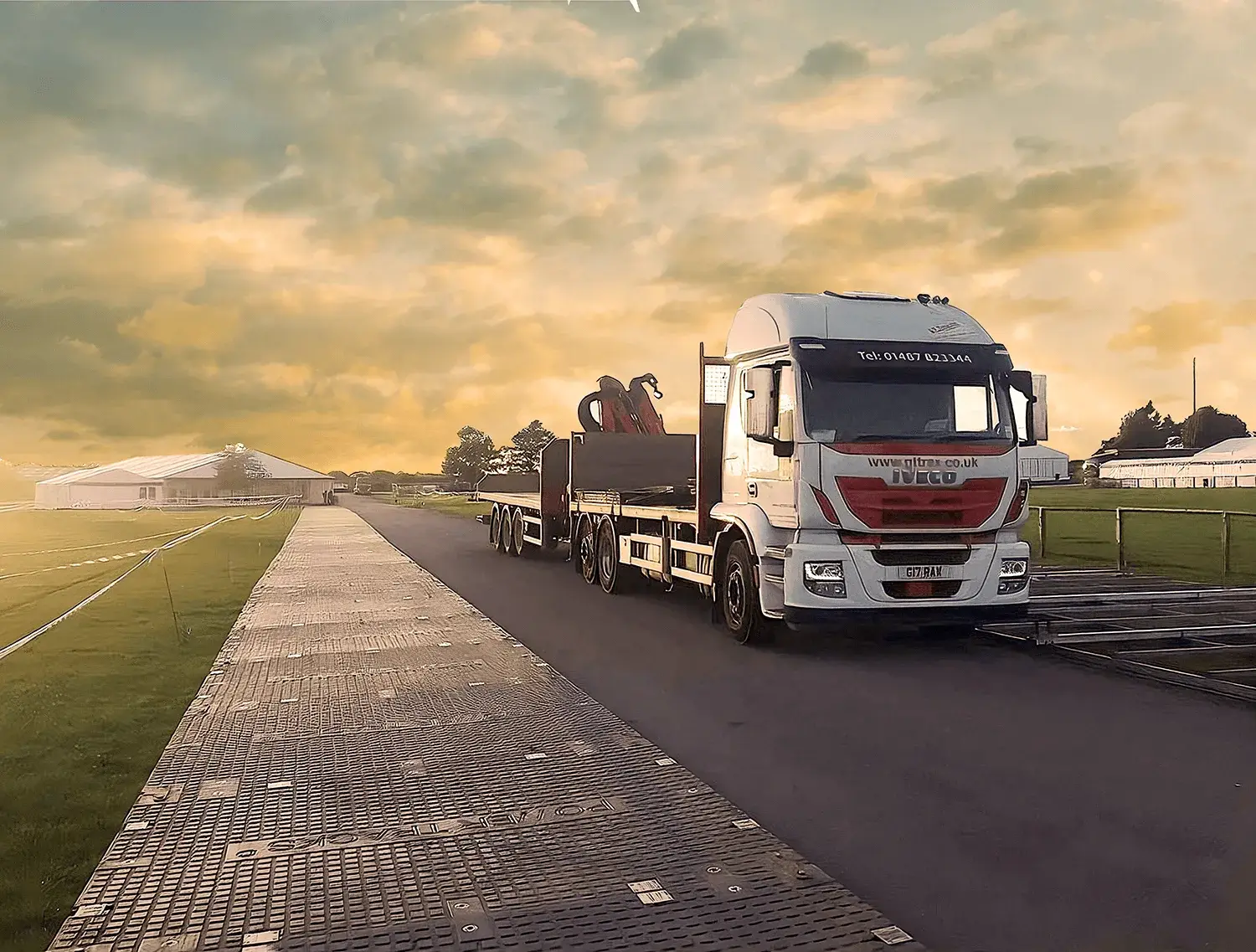 White truck with flatbed trailer and crane on road, beside temporary PowerTrax 60 mats. Large white marquees in background.