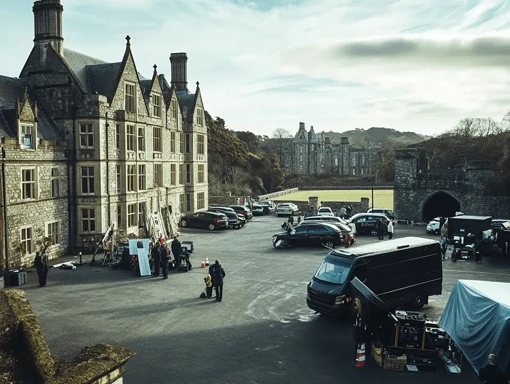 Film crew with equipment in a paved courtyard by a stone mansion. Another castle, hills, and cars in the background under a cloudy sky.