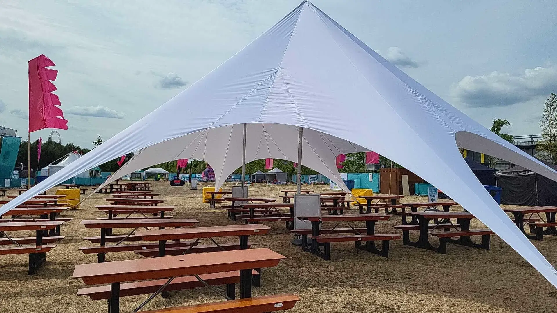 A large white star-shaped StarShade canopy shades rows of empty wooden picnic tables on a dry outdoor field with flags and event structures.