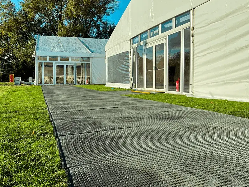 A grey textured EuroMats pathway on grass leads to two white event tents with glass entrances under a blue sky.